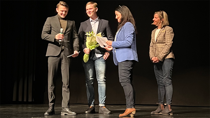 Bild på fyra personer i en intervjusituation på en scen med svart bakgrund. En person håller i en mikrofon. En annan person håller i en blombukett. Bild på fyra personer i en intervjusituation på en scen med svart bakgrund. En person håller i en mikrofon. En annan person håller i en blombukett.
